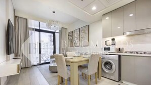 A modern kitchen with a built-in washing machine, a dining area with a table and chairs, and a large window with curtains. - 520 Bloomfields 1BR Azizi Riviera (Dubai)