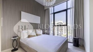 A modern bedroom with a large bed, a small side table, a chandelier, and a view through large windows. - 520 Bloomfields 1BR Azizi Riviera (Dubai)