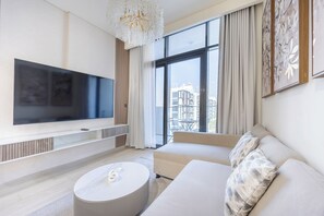 A modern living room with a flat-screen TV, a sofa, a coffee table, and a large window offering a city view. - Bloomfields 1BR in Azizi Riviera 33 - Meydan (Dubai)