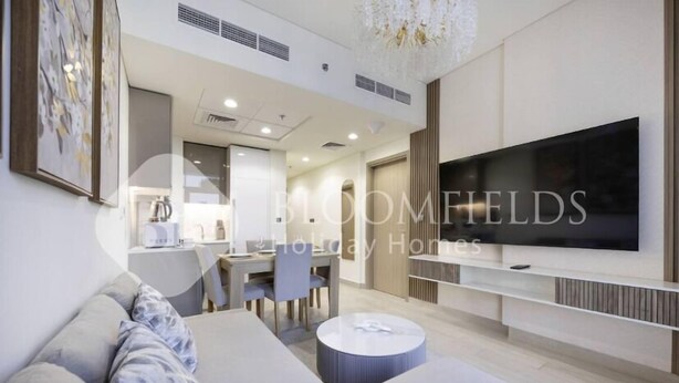 A modern living room with a flat-screen TV, a sofa, a dining area, and a chandelier. - 520 Bloomfields 1BR Azizi Riviera (Dubai)