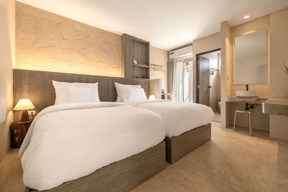 A modern hotel room with a large bed, a bedside table with a lamp, a built-in shelf, and a bathroom with a sink and mirror.