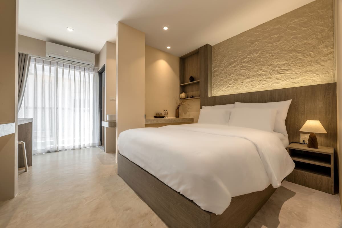 A modern hotel room with a large bed, a nightstand, a wall-mounted shelf, and a window with sheer curtains.