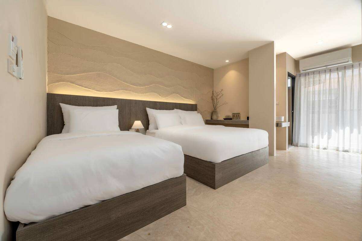 A modern hotel room with two beds, a large wall-mounted artwork, and a large window with sheer curtains.