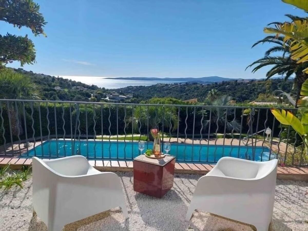 A poolside area with two white chairs and a small table with a vase and glasses, overlooking a pool and a scenic view of hills and water.