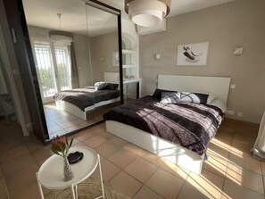Ein Schlafzimmer mit einem großen Spiegel, einem Bett mit dunkel gemustertem Überwurf, einem kleinen runden Tisch mit einer Vase und einem Fenster mit Jalousien.