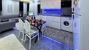 Una sala de estar moderna con sofá, mesa de comedor y sillas. Cuenta con una zona de cocina, lavadora y televisión.