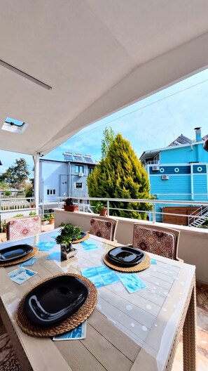 An outdoor dining area with a table set for four, surrounded by chairs, and overlooking a residential area with houses and a large tree.
