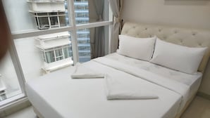 Un lit bien fait, avec du linge de lit blanc, dans une chambre d’hôtel dotée de grandes fenêtres et offrant une vue sur les bâtiments environnants.