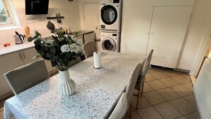 Un comedor con una mesa dispuesta para cuatro personas, sillas alrededor, un florero con flores y una cocina al fondo con lavadora.