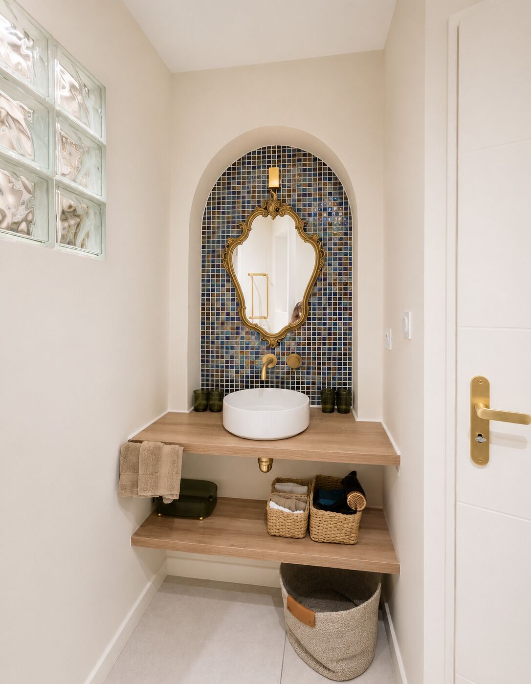 Ein Badezimmer mit einem weißen Waschbecken, einem Spiegel mit aufwändigem Rahmen, einer gefliesten Rückwand und einem Holzregal mit Körben.