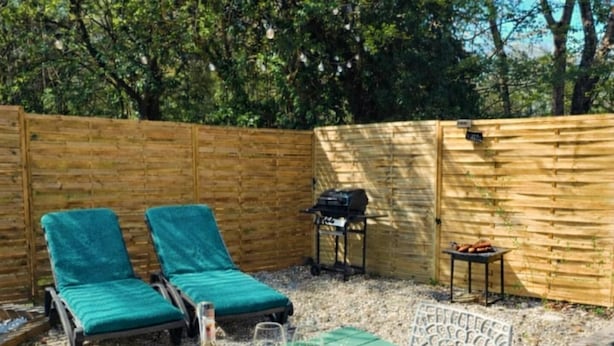 Un patio con tavolo apparecchiato per un pasto, due poltrone lounge, un barbecue e un recinto in legno.