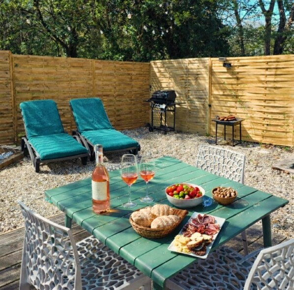 Une terrasse avec une table dressée pour un repas, deux chaises longues, un barbecue et une clôture en bois.