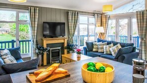 Ein Wohnzimmer mit Kamin, Fernseher, einer Couch, einem Couchtisch mit Brot und einer Obstschale, sowie großen Fenstern mit Blick ins Freie.
