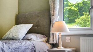 Ein Schlafzimmer mit einem Bett, einem Nachttisch, einer Lampe, einem Fenster mit Blick auf Grünflächen und einem Heizkörper.