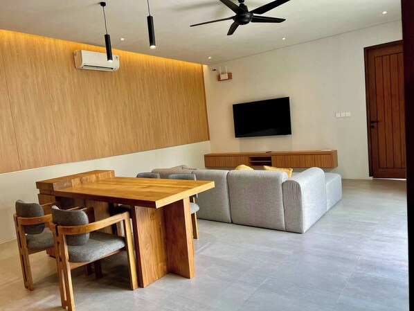 A modern living room with a wooden dining table, grey sofa, and a flat-screen TV mounted on the wall. - Guru Darma Villa Newly Built September 2025 (Taro)
