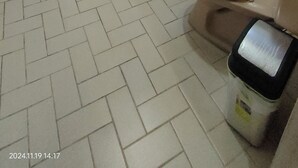 Une petite salle de bain carrelée, équipée de toilettes, d’un lavabo et d’une douche. On y trouve un seau rouge et une grande poubelle bleue.