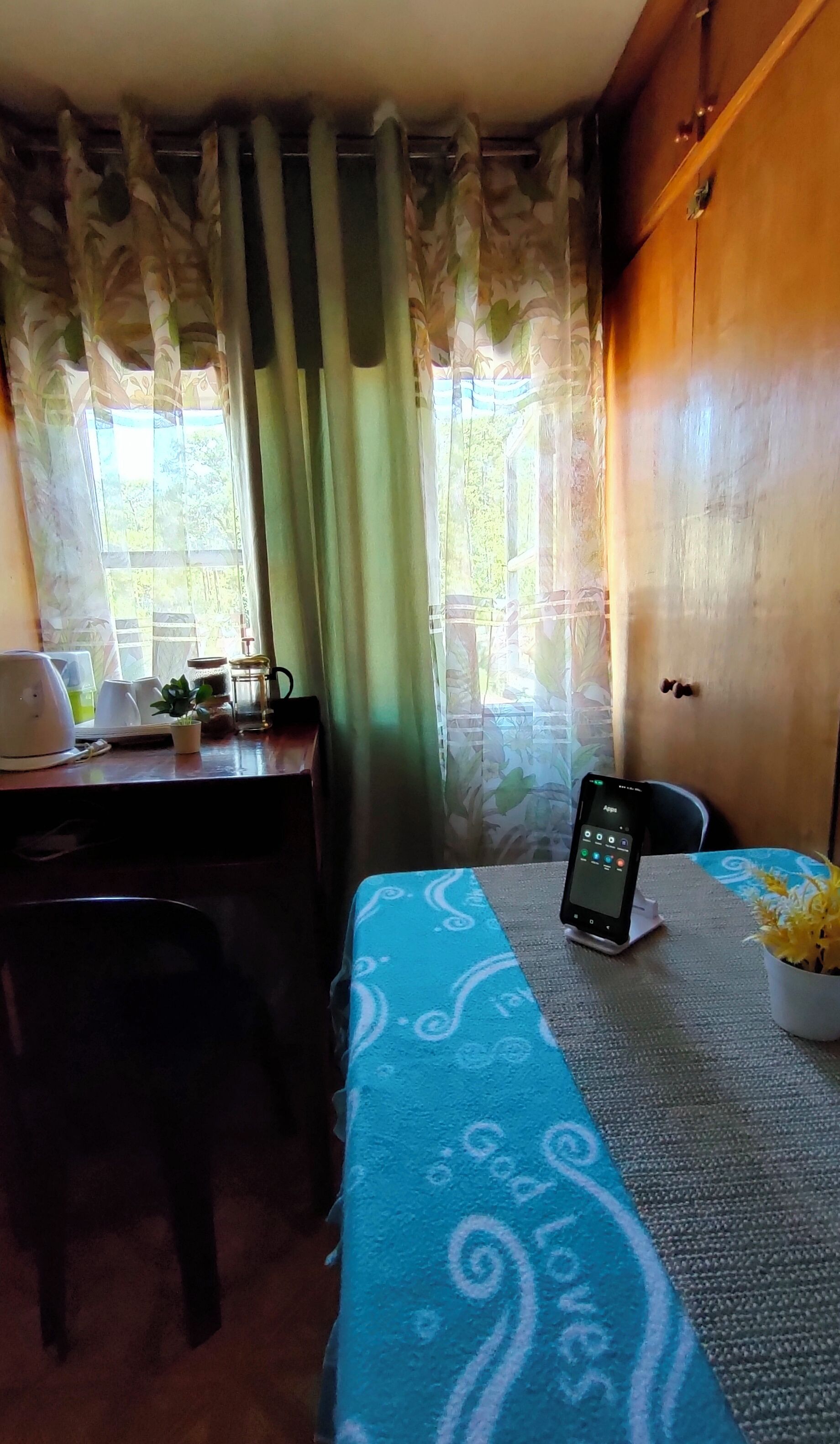A room with a desk, a chair, a bed with a blue patterned bedspread, a smartphone on the bed, a potted plant, and a window with curtains.