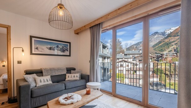Ein modernes Wohnzimmer mit grauem Sofa, einem Holztisch und einer großen Glasschiebetür, die auf einen Balkon mit Bergblick führt.