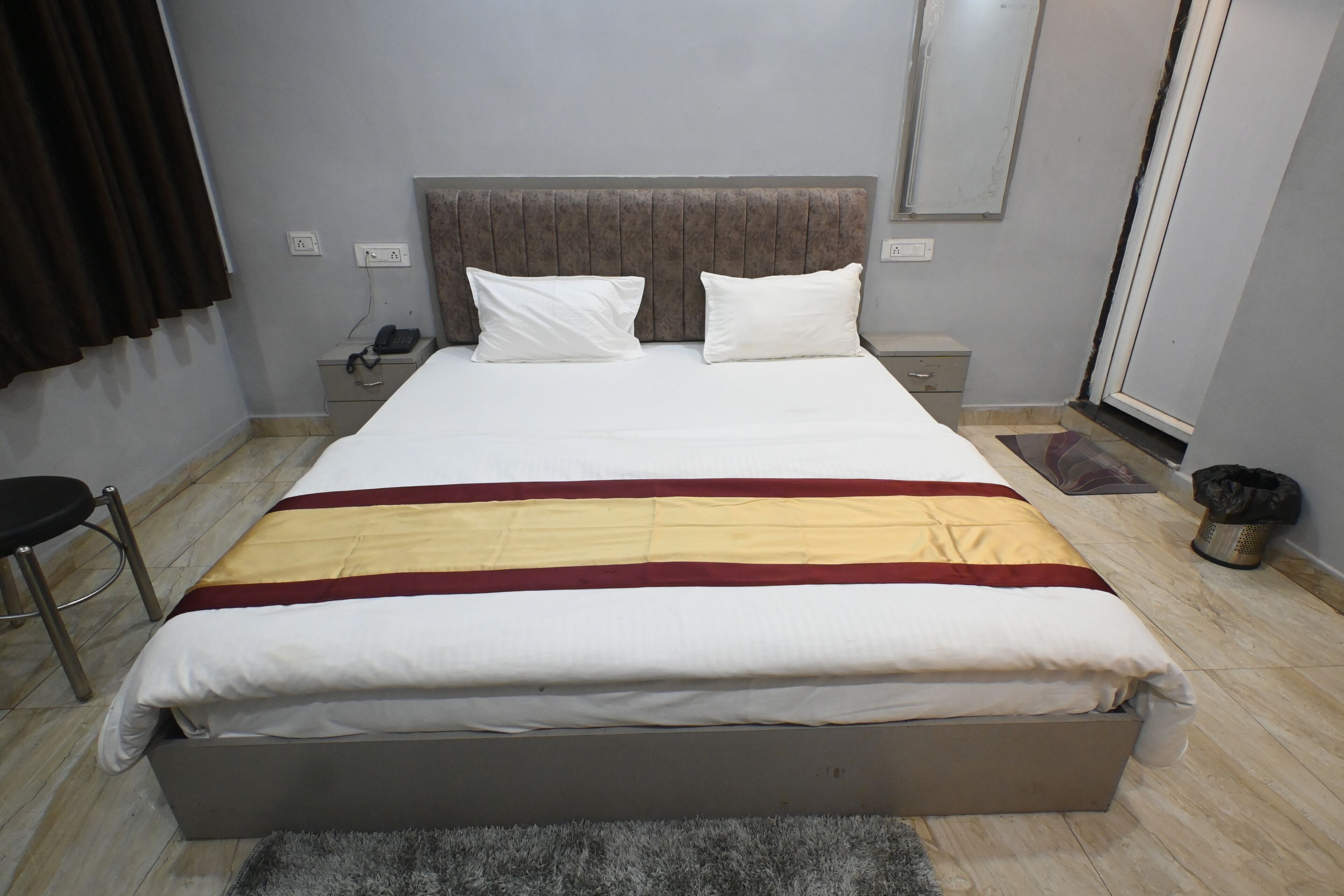 A neatly made bed with a grey headboard, a bedside table with a phone, and a small rug on the floor.