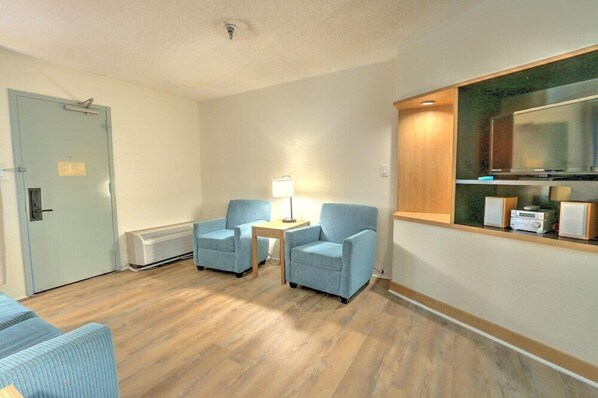 A hotel room with a wooden floor, two blue armchairs, a small table with a lamp, a TV, and a minibar. - Hotel Carolina a Days Inn by Wyndham (Hilton Head Island)