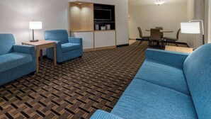 Una habitación de hotel con alfombra estampada, un sofá azul, una mesita con lámpara, un mueble para televisión y un comedor con sillas y una mesa.