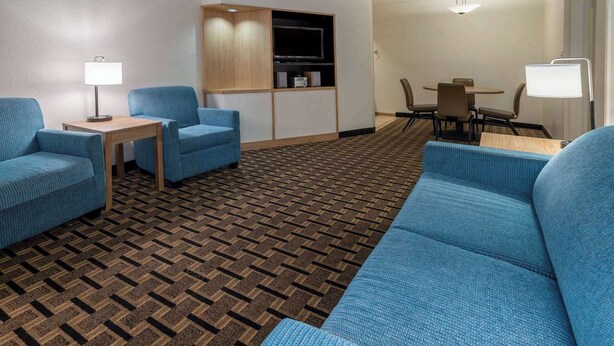 Una habitación de hotel con alfombra estampada, un sofá azul, una mesita con lámpara, un mueble para televisión y un comedor con sillas y una mesa.