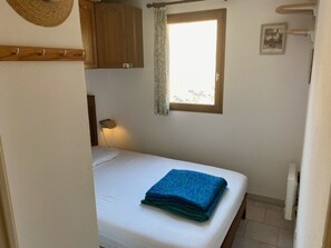 Ein kleines, ordentlich gemachtes Bett mit blauer Decke in einem kompakten Zimmer mit Fenster, Holzschränken und einer wandmontierten Lampe.