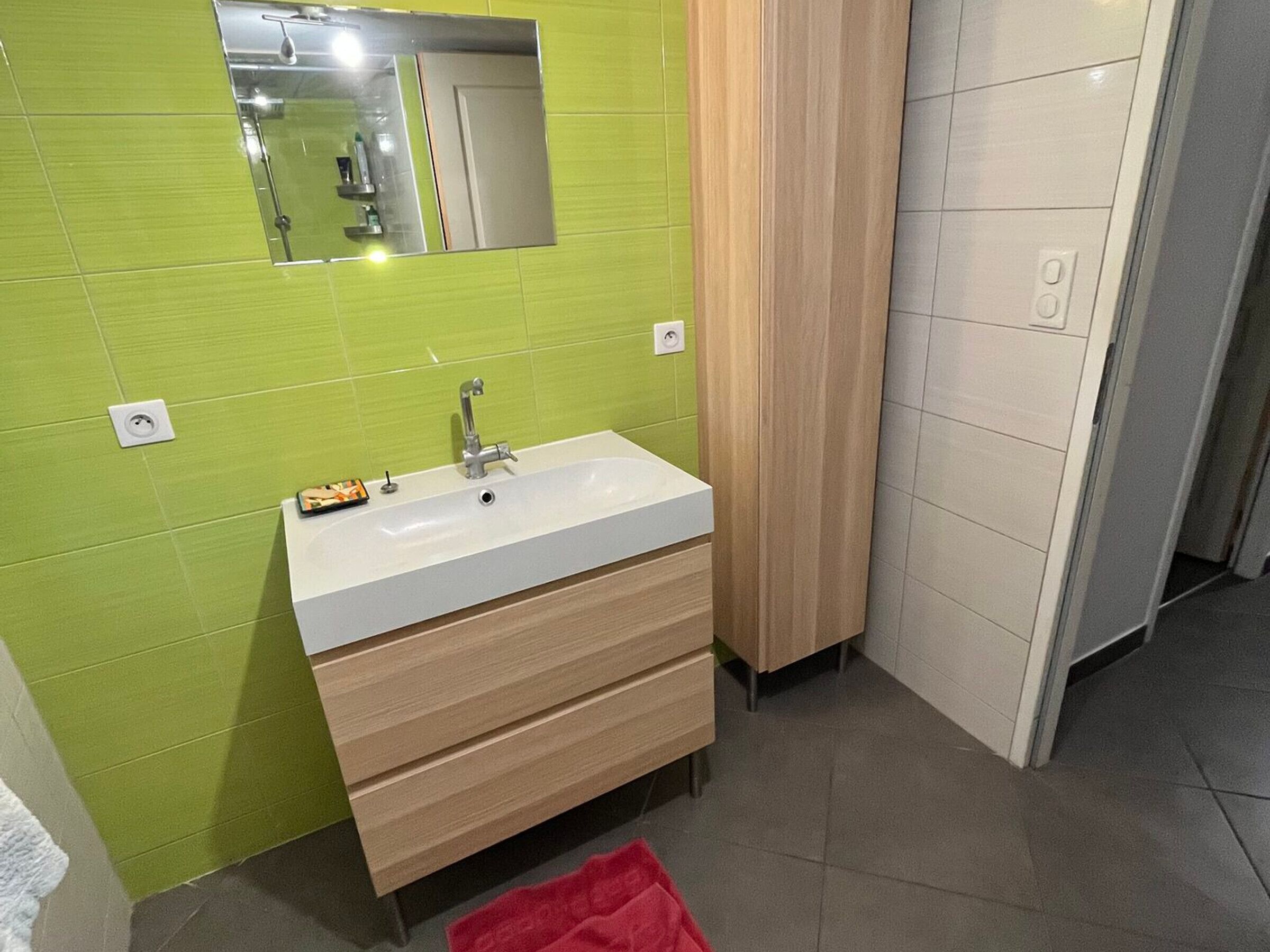 Un baño con lavamanos blanco, paredes con azulejos verdes, tocador de madera y una toalla roja en el suelo.