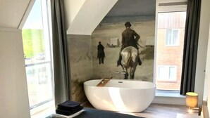 Une chambre à coucher moderne avec une grande baignoire, un lit doté d’une tête de lit sombre et une grande fenêtre.