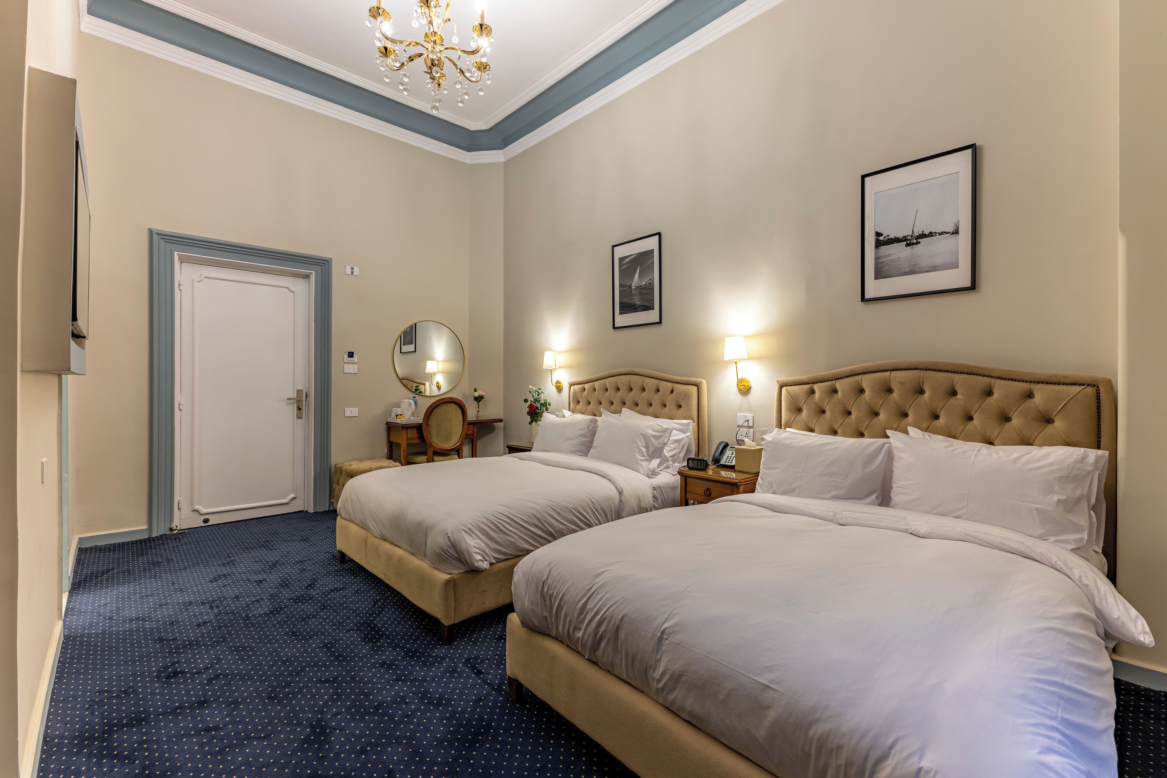 Habitación de hotel con dos camas, una araña, un escritorio con silla y cuadros en las paredes.