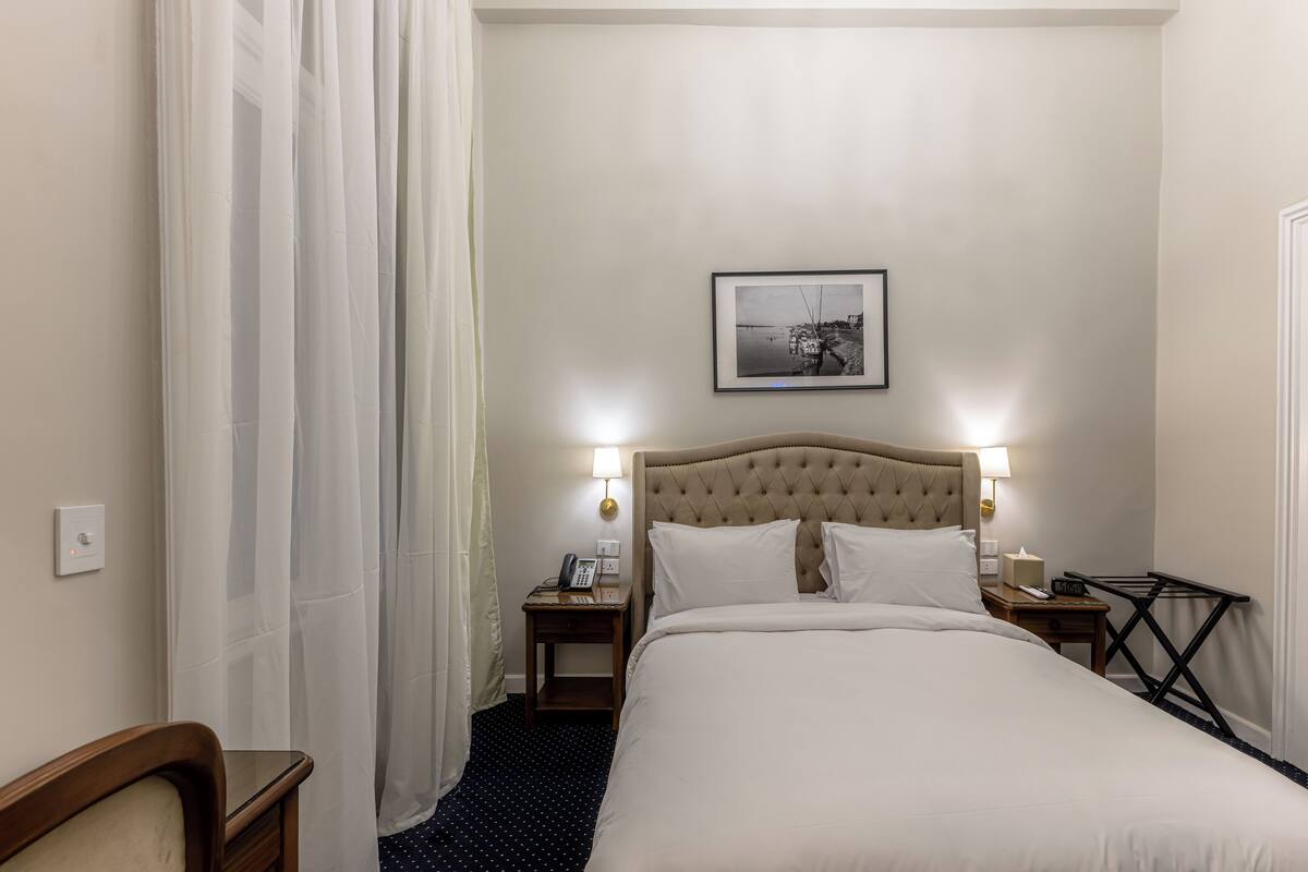 Habitació d'hotel amb un llit gran, dues taules de nit amb làmpades, un quadre al mur i una cortina a la finestra.