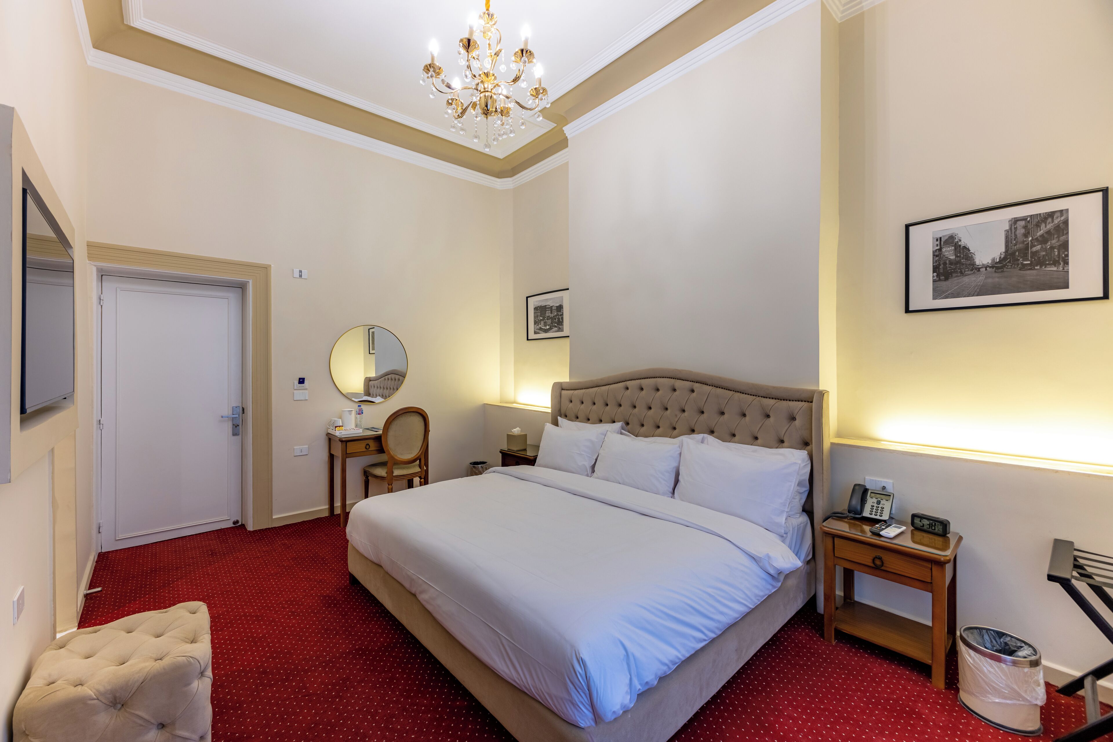 A hotel room with a large bed, a chandelier, a bedside table with a phone, a chair, and a small table with a lamp.