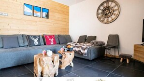 A modern living room with a grey sofa, wooden wall paneling, a large clock, and a rustic coffee table. - Apartment in Bonneval Near Vallonnet Lift (Bonneval-sur-Arc)