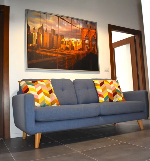 A modern living room with a blue sofa, colorful geometric pillows, and a large cityscape painting on the wall.