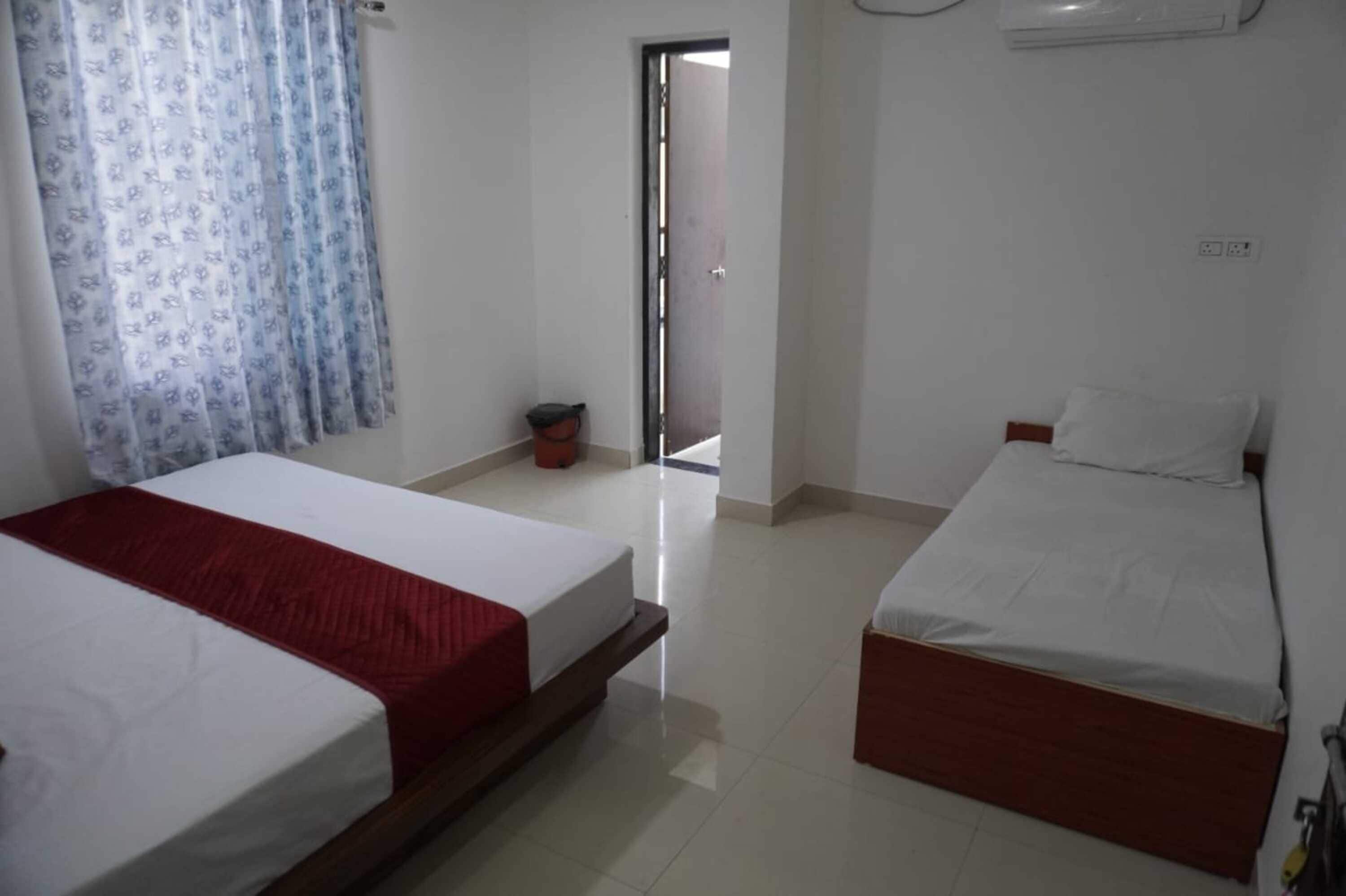 Una camera d'albergo con due letti, una coperta rossa su un letto e una bianca sull'altro, una finestra con tende blu e una porta che dà su un balcone.