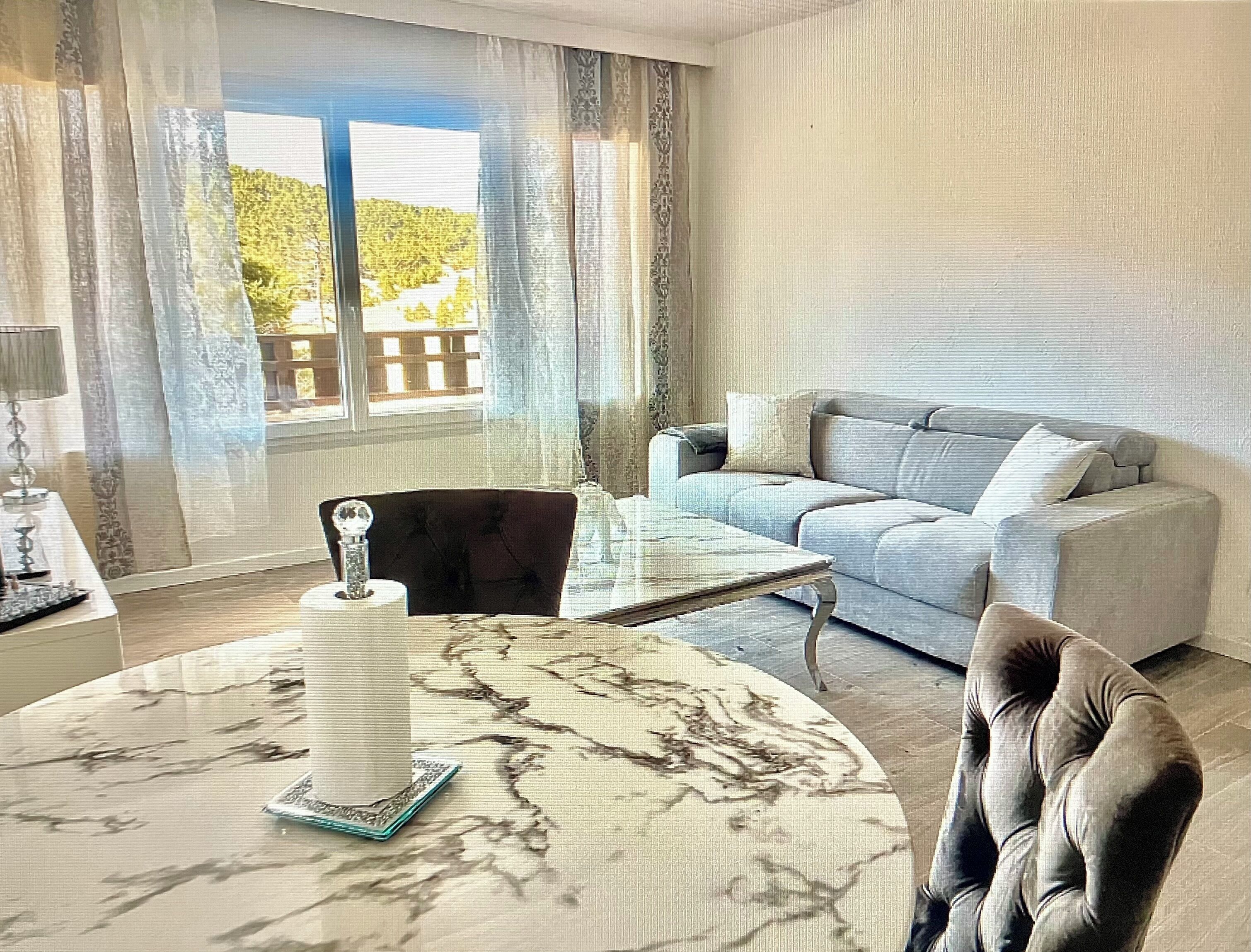 Een woonkamer met een bank, een salontafel en een stoel. Er is een raam met gordijnen, een lamp en een decoratief object op de tafel.