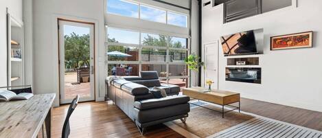 A spacious living area with a large sofa, wooden flooring, and a view of the outdoors through large windows.