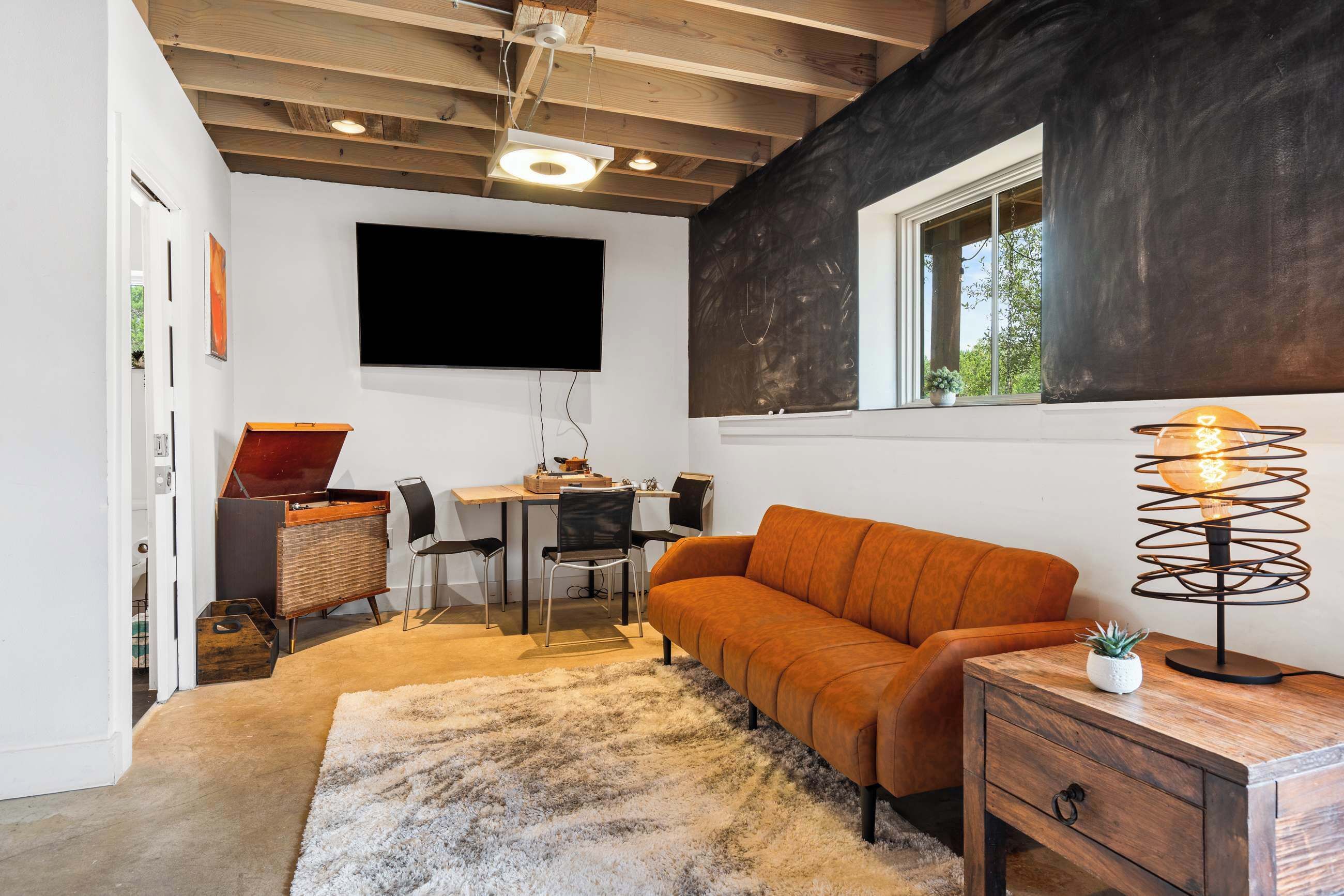 A modern living room with a wooden ceiling, a flat-screen TV mounted on the wall, a wooden side table with a lamp, a sofa, and a desk with a chair.