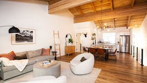 Ein modernes Wohnzimmer mit einer Couch, einem Sessel und einem Couchtisch, ein Essbereich mit Stühlen, eine Küche mit Geräten und einer Holzbalkendecke.