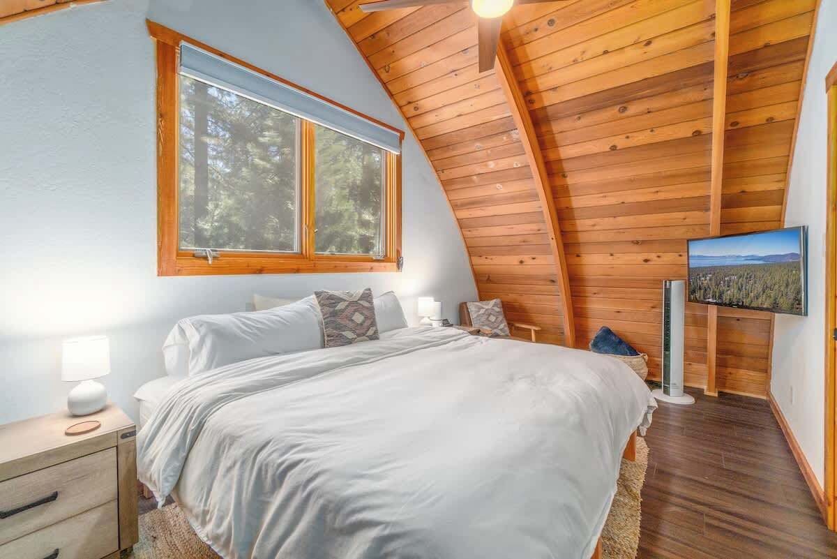 Een slaapkamer met een houten plafond, een groot bed, een nachtkastje, een lamp, een televisie en een raam met uitzicht op bomen.