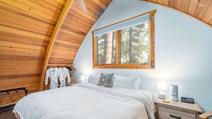 A bedroom with a wooden ceiling, a large bed, a nightstand, a lamp, and a window with a view of trees. - The Canoe Cabin in Incline Village (Incline Village)