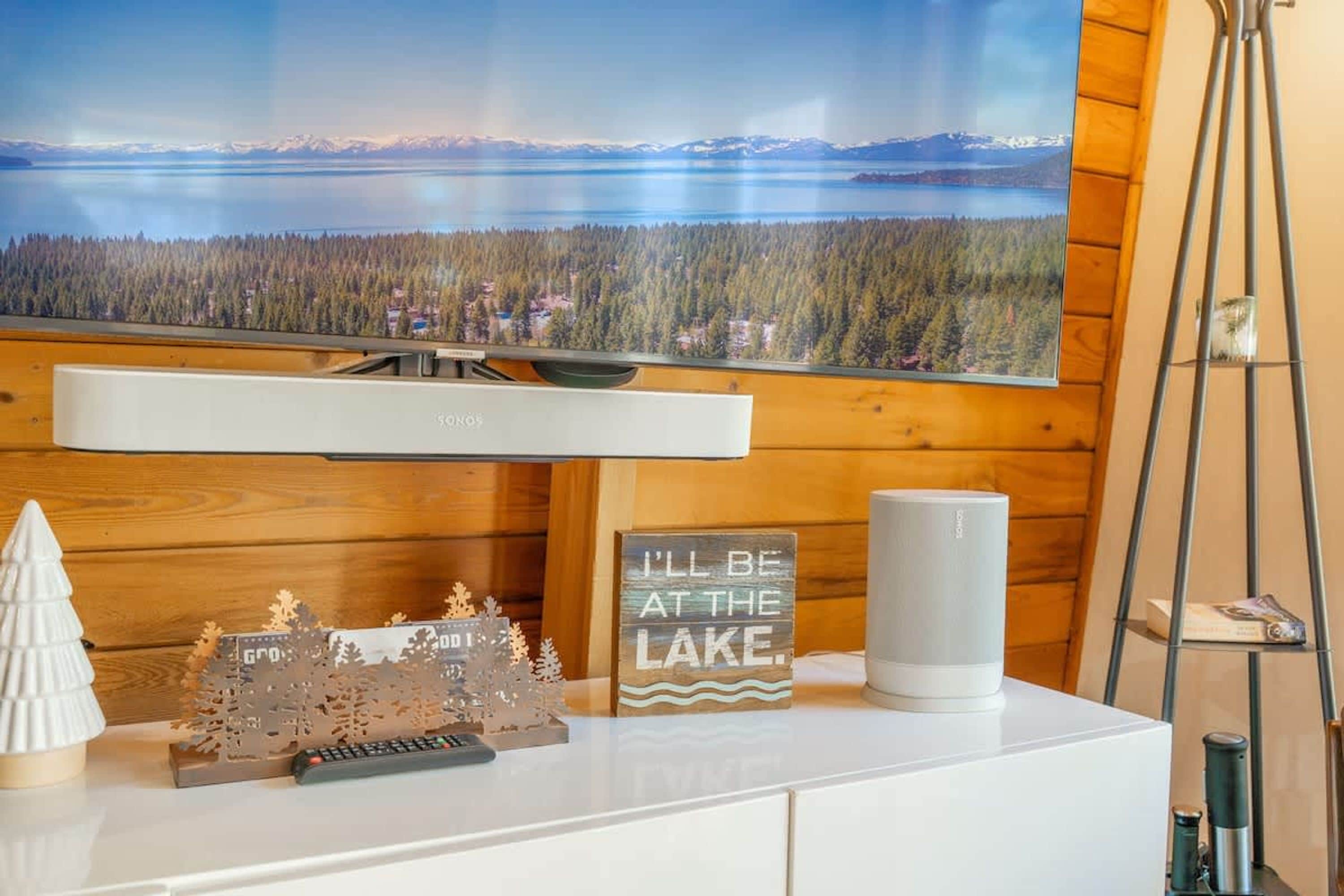 A room with a large flat-screen TV mounted on a wooden wall, a white entertainment unit with a remote control, a decorative sign, and a white speaker.