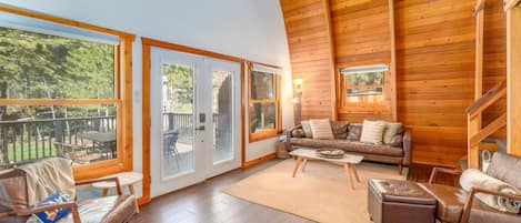 A spacious living area with a high ceiling, wooden walls, and large windows offering a view of the outdoors.