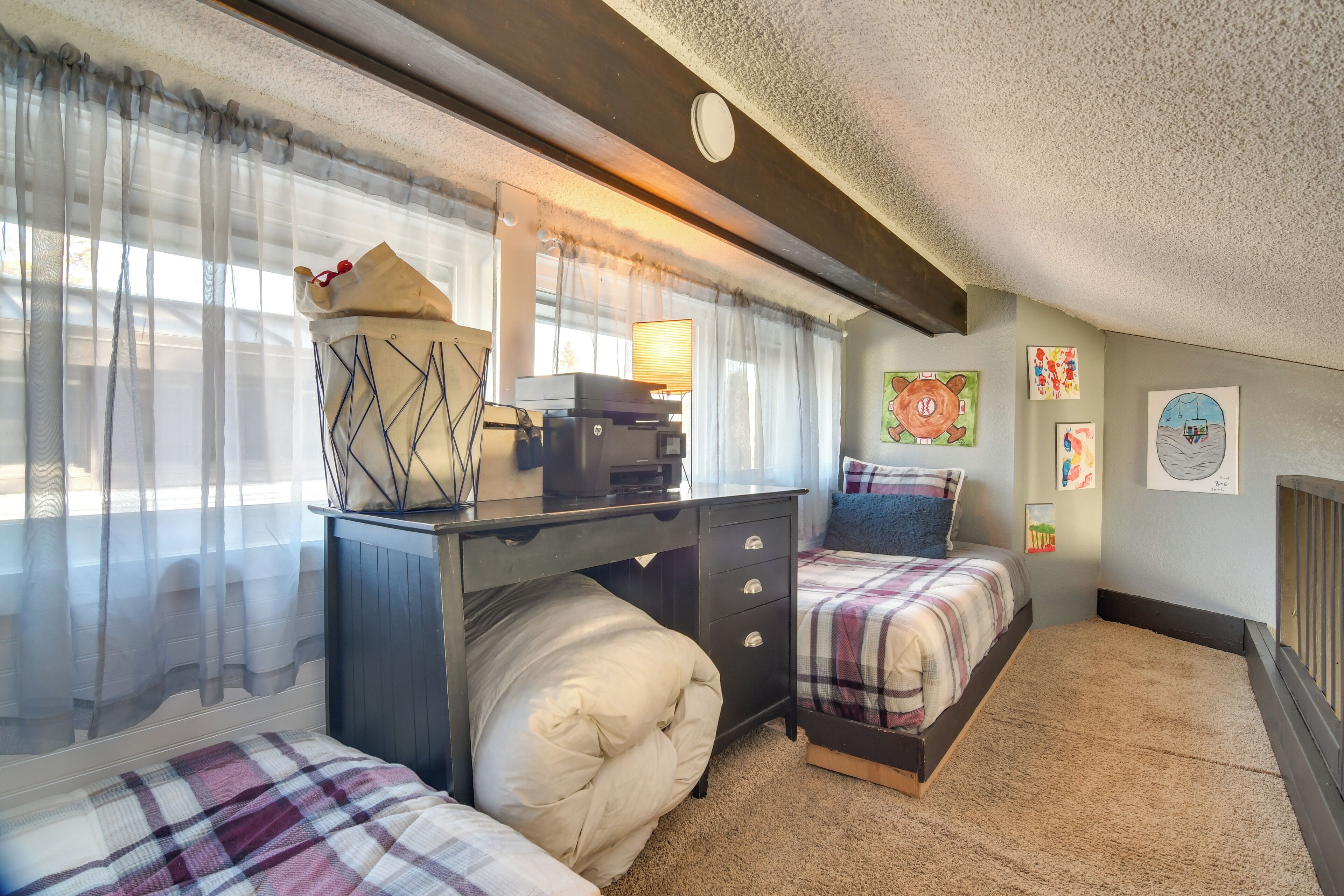 A cozy bedroom with two beds, a desk with a computer, and a window with sheer curtains.