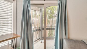 Habitación con puerta corrediza de vidrio que da a un espacio exterior con vistas a árboles y un edificio. En la habitación hay una mesa de madera y un banco.