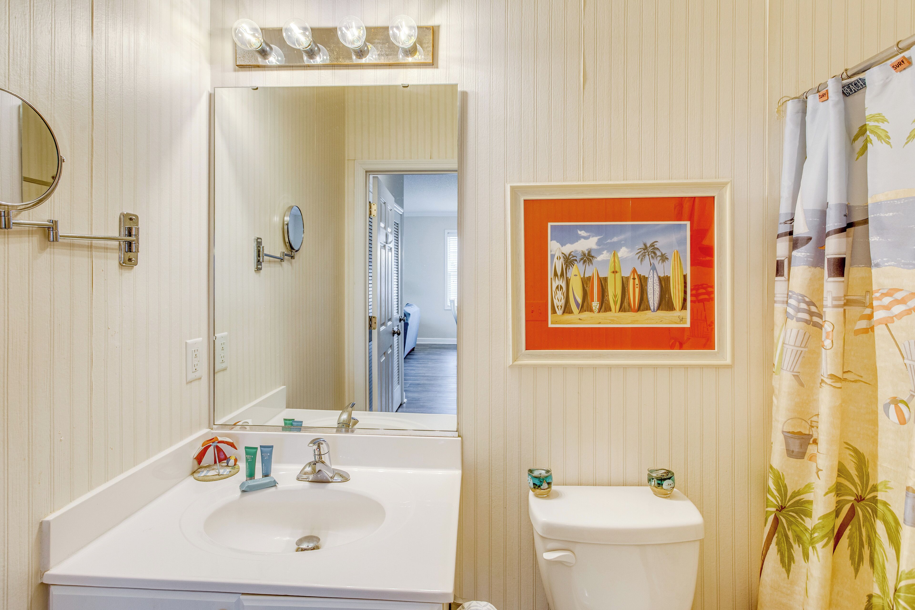 A bathroom with a white sink, mirror, and toilet. There is a framed picture on the wall and a shower curtain with a tropical design.