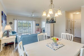 A dining area with a white table, chairs, and a bowl of fruit. There is a ceiling fan, a window with curtains, and a painting on the wall. - Golf & Relax! Resort-style Condo in Myrtle Beach (North Myrtle Beach)
