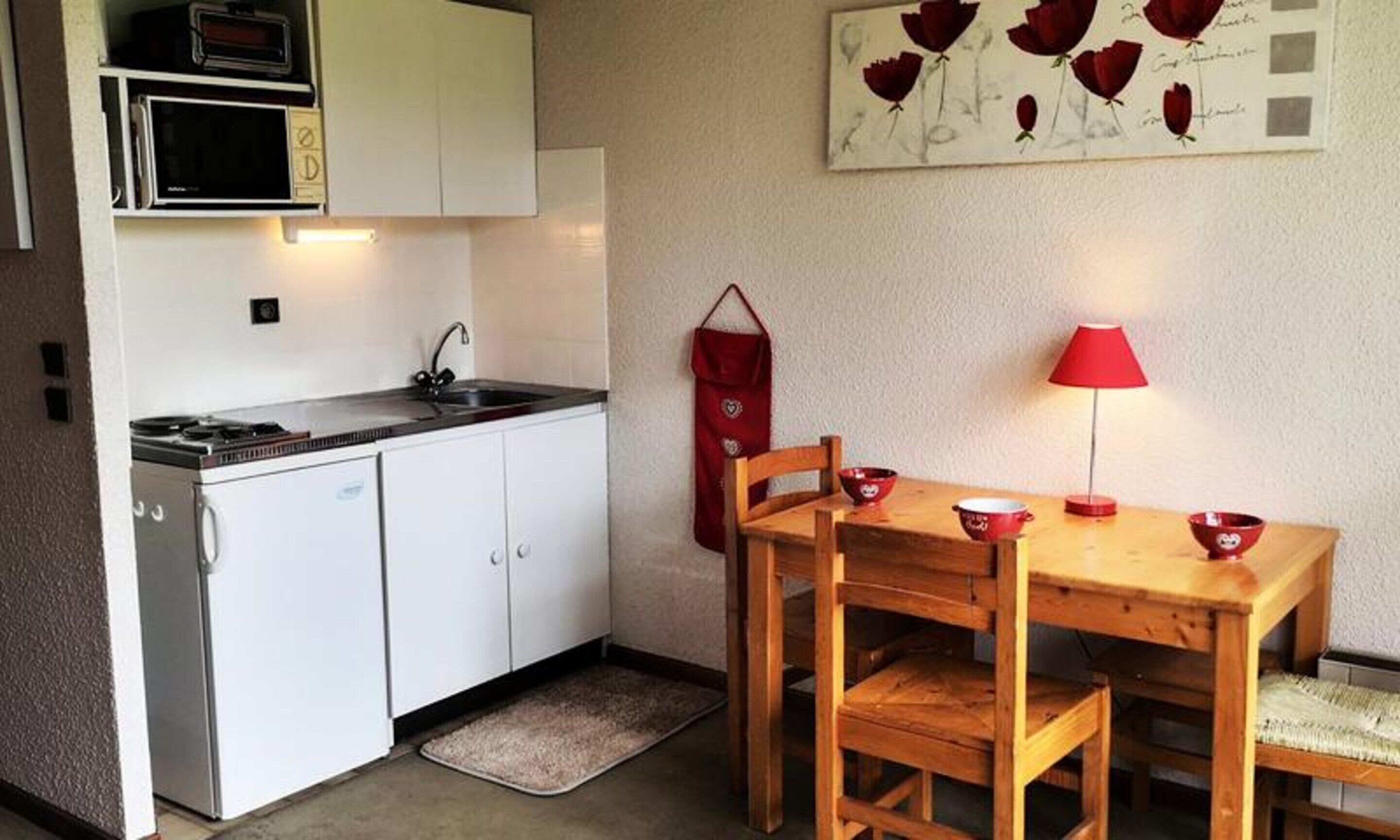 Una cocina compacta con alacenas blancas, estufa y microondas. Hay una mesa de madera con sillas y una lámpara de mesa roja. Una tela roja cuelga de la pared, y un tapete pequeño está en el suelo.