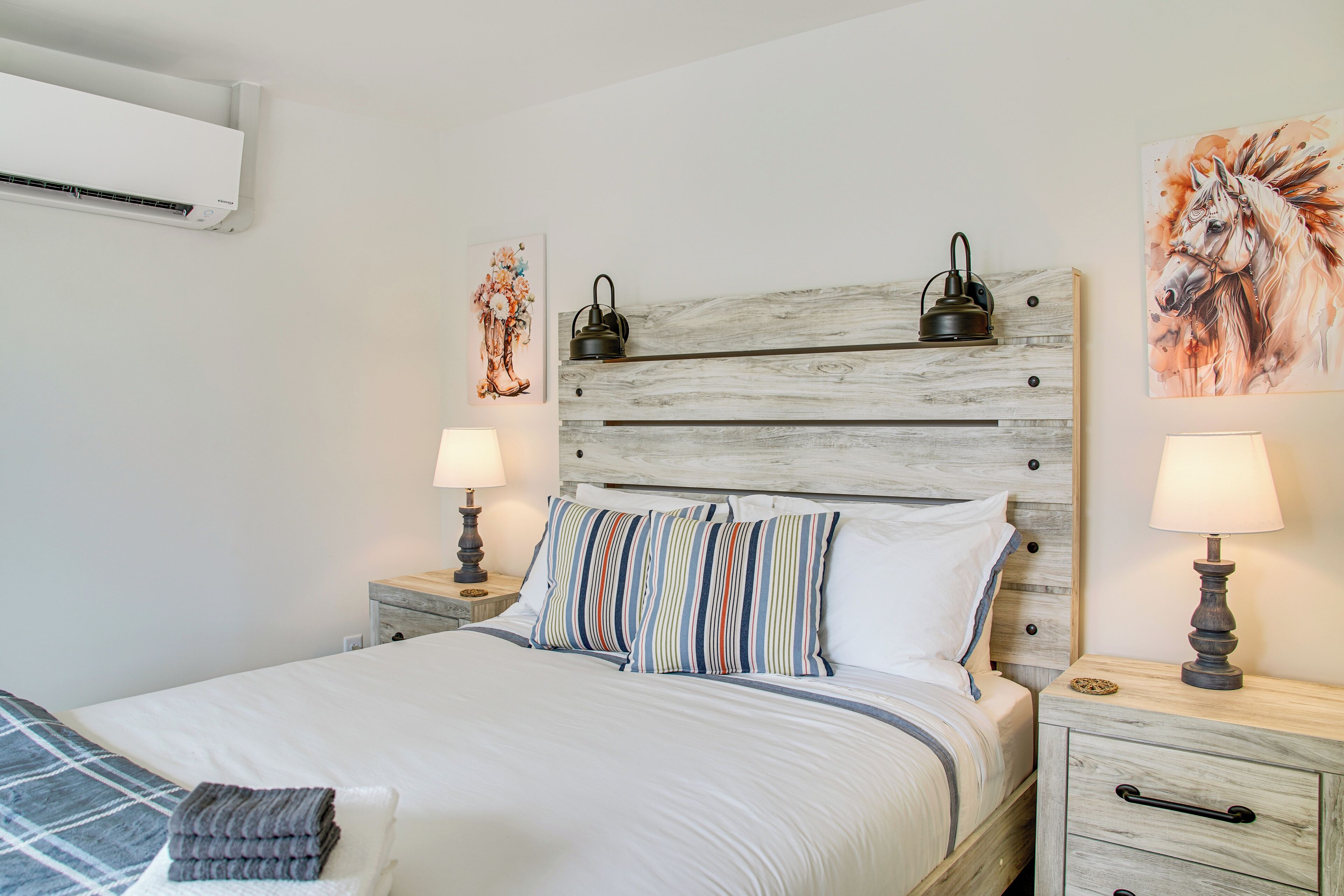 A neatly made bed with striped pillows, a wooden headboard, bedside lamps, and a wall-mounted air conditioner.