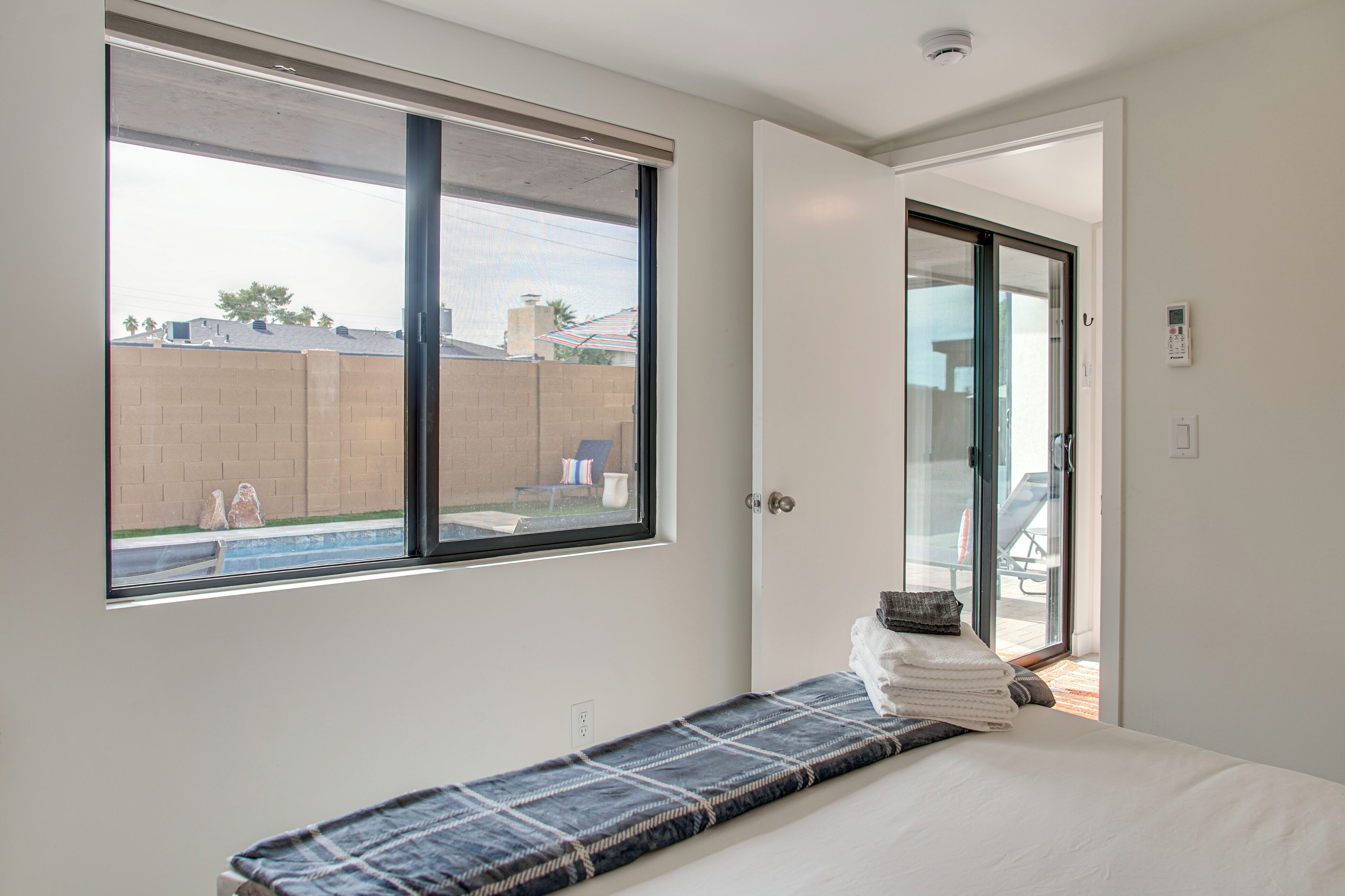 A bedroom with a large window, a bed with a blue and white striped bedspread, and a view of a backyard with a pool and patio furniture.